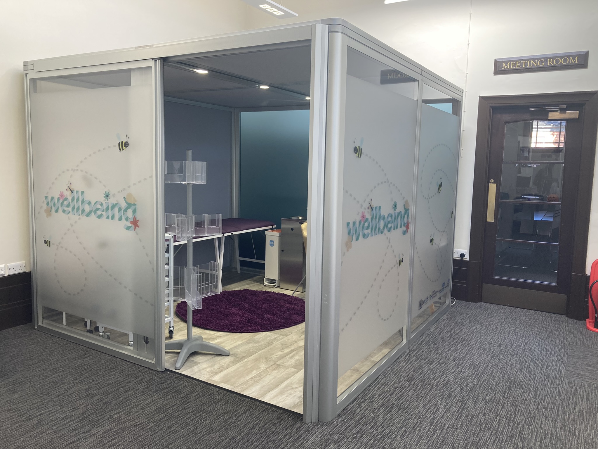 A small enclosed room within a larger room. It is enclosed using frosted panels with the word 'wellbeing' printed on the side. Inside the smaller room, there is a clinical bed, a carpet and some medical trays,