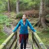 This image shows a woman standing on a wooden bridge in the middle of a forest. She is smiling.