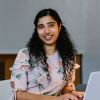 This is a photo of Ashly, a Masters student studying MSc Biopharmaceutical Development. She is sitting down in front of a table working on a laptop. There is a lamp next to her which is turned on. She is looking at the camera.