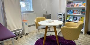 A maternity service within a community hub in Leeds. A small round table and two yellow chairs are in the centre of the room. Also in the room are white shelves, a moveable screen and sink.