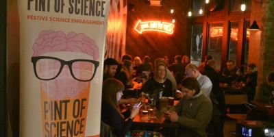 This image shows a room full of people laughing and talking. In the foreground there is a pull up banner advertising Pint of Science, the world's largest festival of public science talks.