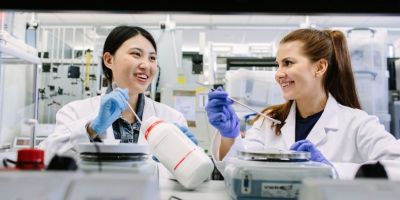 Two female scientists in white lab coats smile at each other.