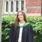 This picture shows a woman standing in front of a red brick building and a hedge. She is wearing black graduation robes with a green collar. She is smiling.