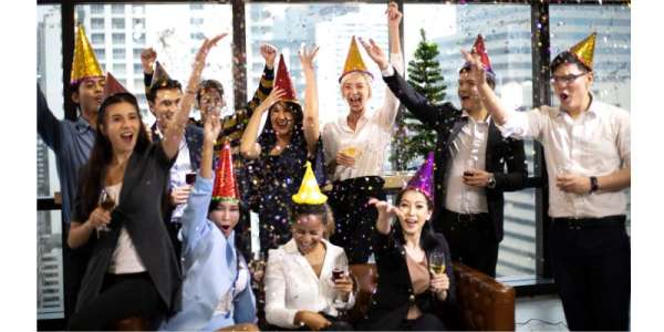 Group of approx. 10 office workers in a high rise office sat in front of a Christmas tree, wearing party hats, holding drinks and throwing glitter.